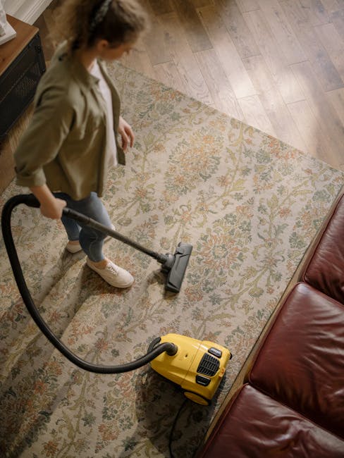 A spacious, empty living room with beige carpet flooring that appears clean and well-maintained. The walls are painted a soft yellow with white trim, and there is a ceiling fan with a light fixture at