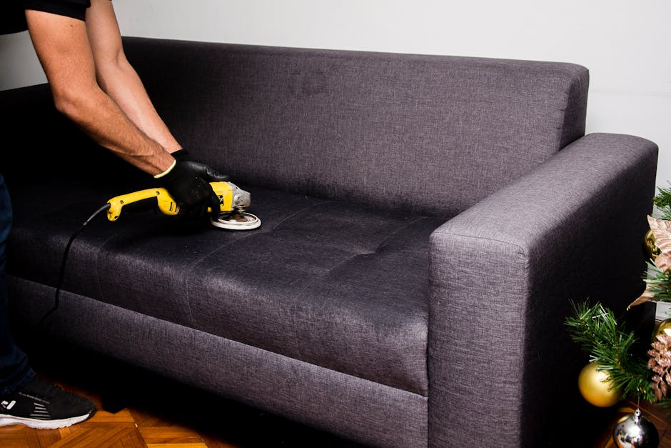 A person wearing black gloves is using a handheld upholstery cleaning tool connected to a yellow and black cleaning device to deep clean a dark grey fabric sofa, situated in a well-lit room with a woo