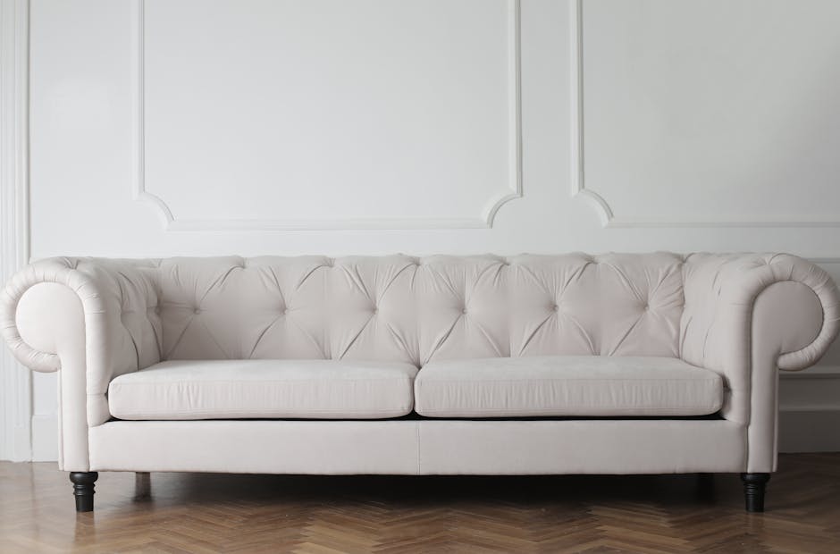A person’s hand holding a vacuum cleaner with a black hose and nozzle, is vacuuming a yellow and white patterned cushion placed on a dark grey fabric sofa in a living room setting. The room has neut